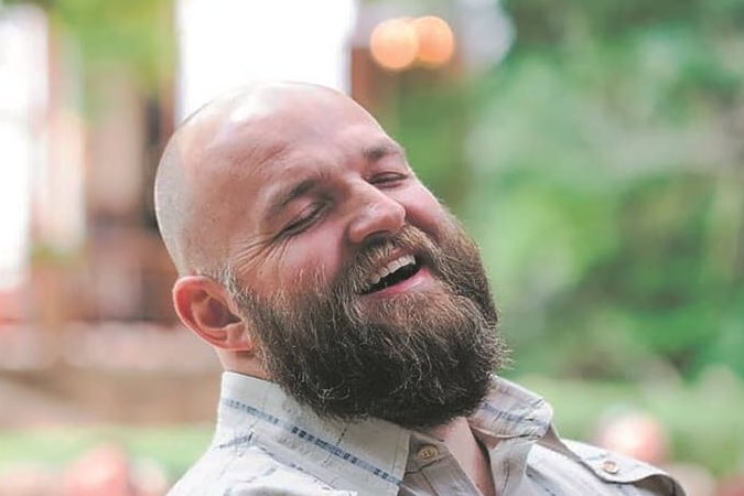 Man with beard and beige shirt laughs with eyes closed. 