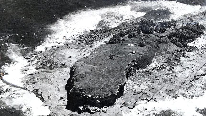 Black and white aerial of a small island showing one structure on the island.