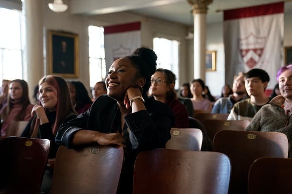 A woman leans on a desk and smiles with a group of students sitting at other desks