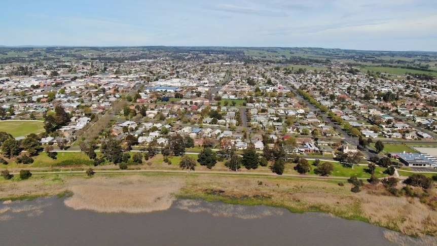 An aerial  photo of a country town built on the edge of a lake, surrounded by green paddocks and hills.