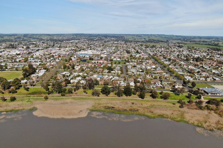 An aerial  photo of a country town built on the edge of a lake, surrounded by green paddocks and hills.