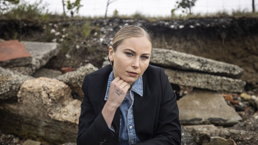 A blonde, fair skinned woman, sits outdoors looking at the camera wearing a blazer