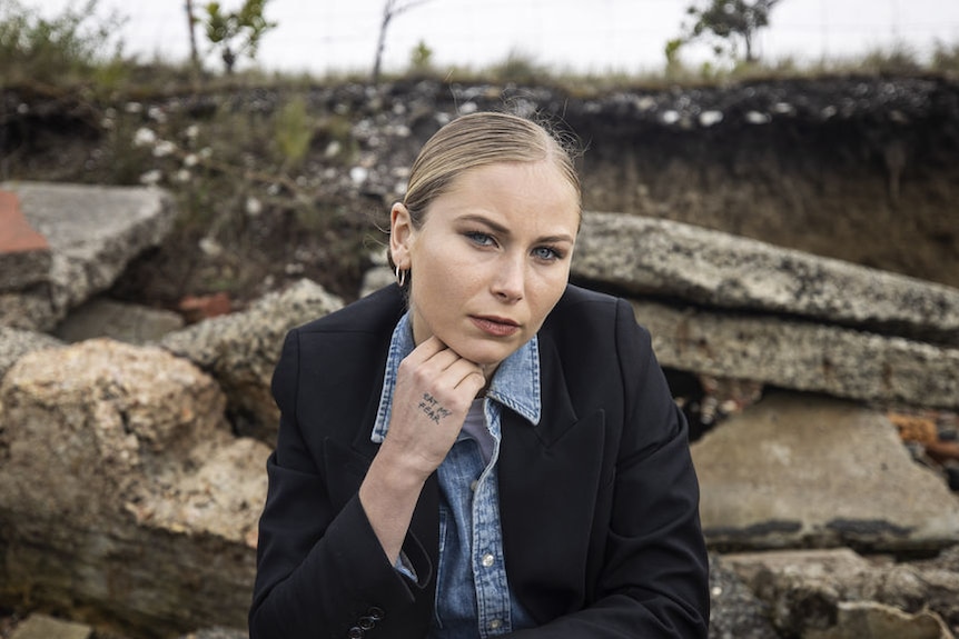 A blonde, fair skinned woman, sits outdoors looking at the camera wearing a blazer