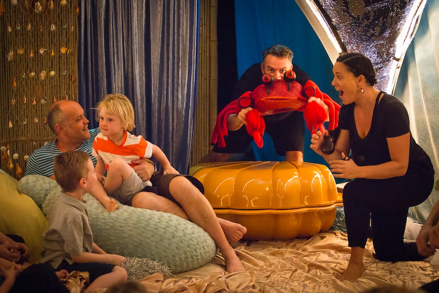 Two small boys sit with their carer on a bean bag being performed to by two performers, one of whom is holding a crab puppet.