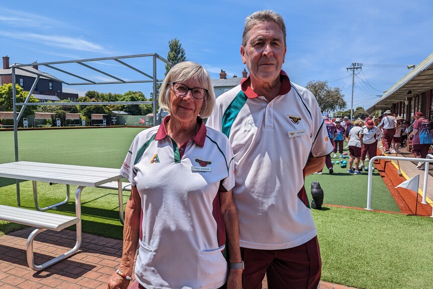 A male and female in their retirement years in their lawn bowls uniform look at the camera with the bowls club in the bckground 