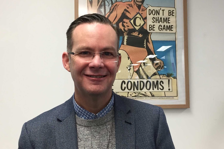 Darryl O'Donnell stands in an office in front of a poster promoting the use of condoms during sex.
