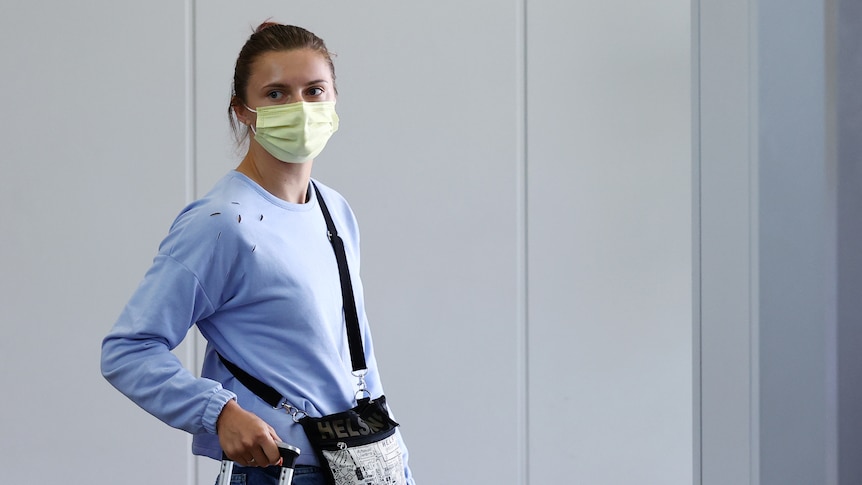 A woman looks sternly while wearing a facemask and carrying luggage at an airport