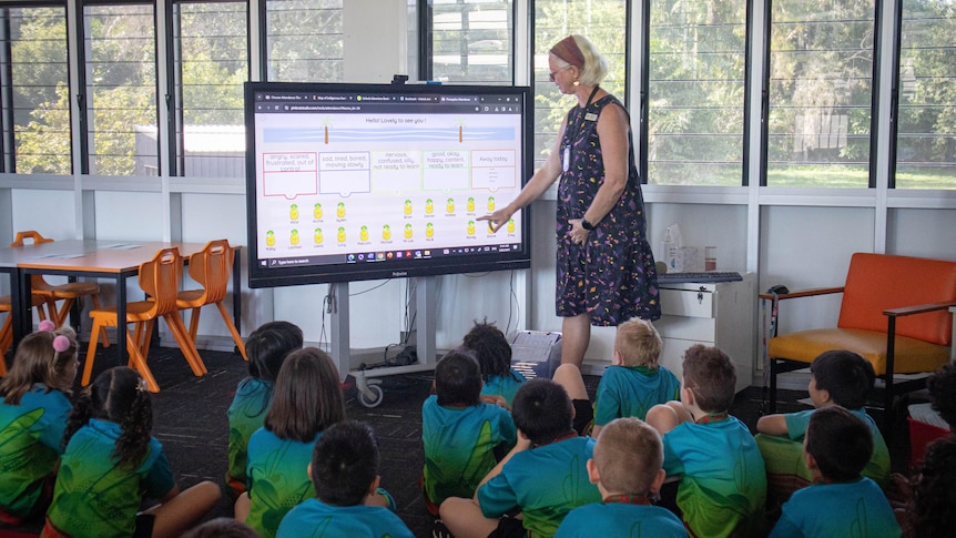 A woman wearing a dress stands in front of a primary school cohort and points to emotions on a screen. 