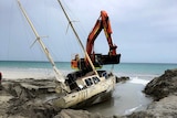 An excavator drags a dilapidated wooden yacht to water through a trench dug into the sand.