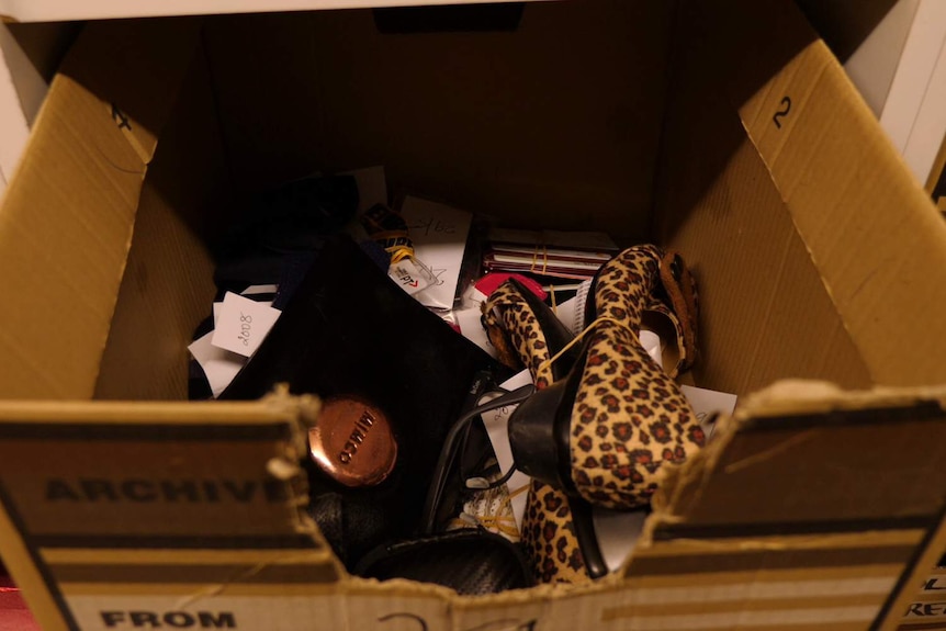 A cardboard box containing a black handbag and leopard print shoes, among a stack of other items