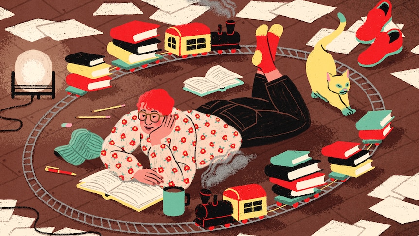 A person lies on their stomach reading a book on the floor, while a model train set bearing more books circles them.