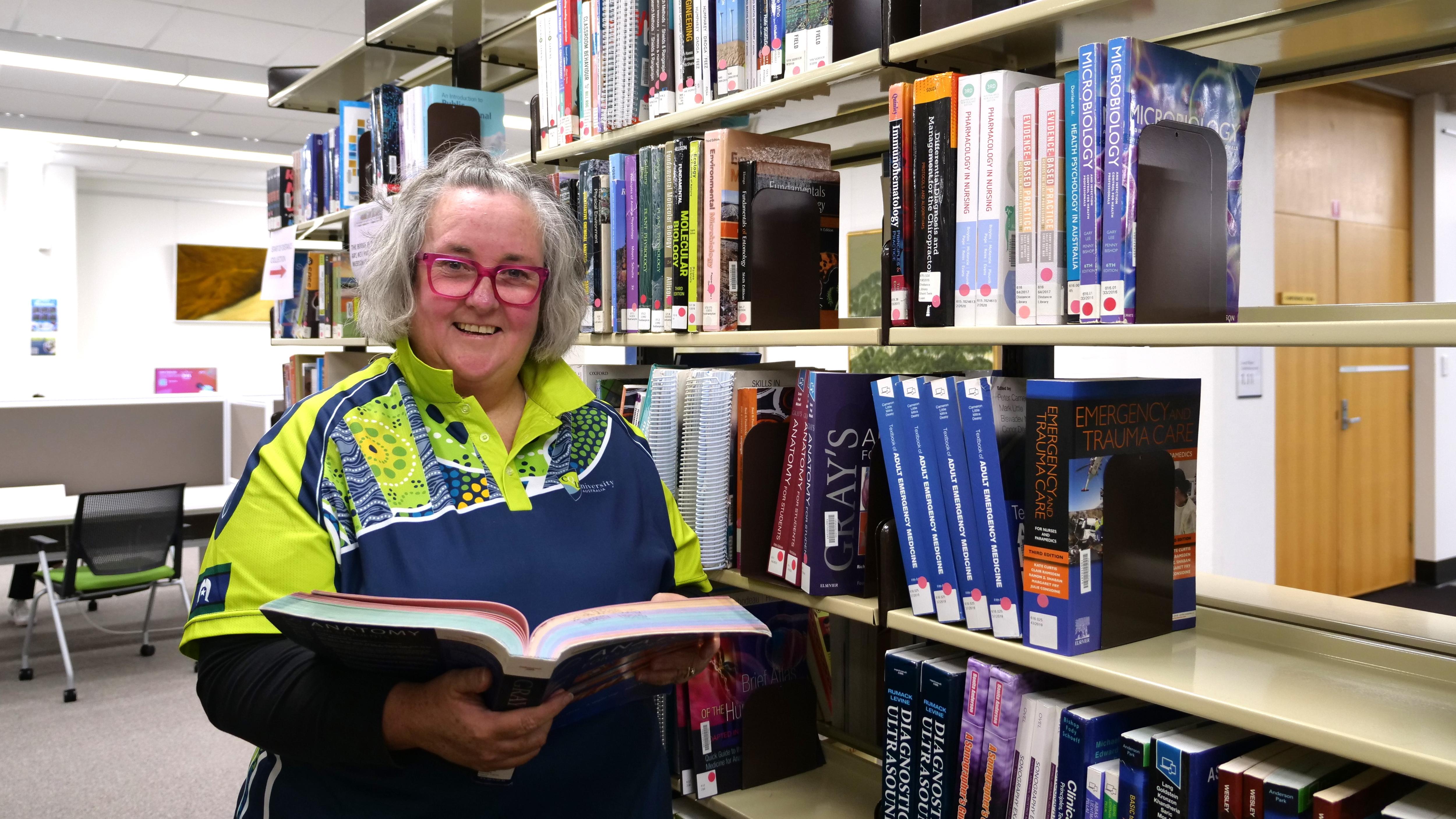 Woman in her photos with bright pink glasses reading a textbook in front of a bookcase. 