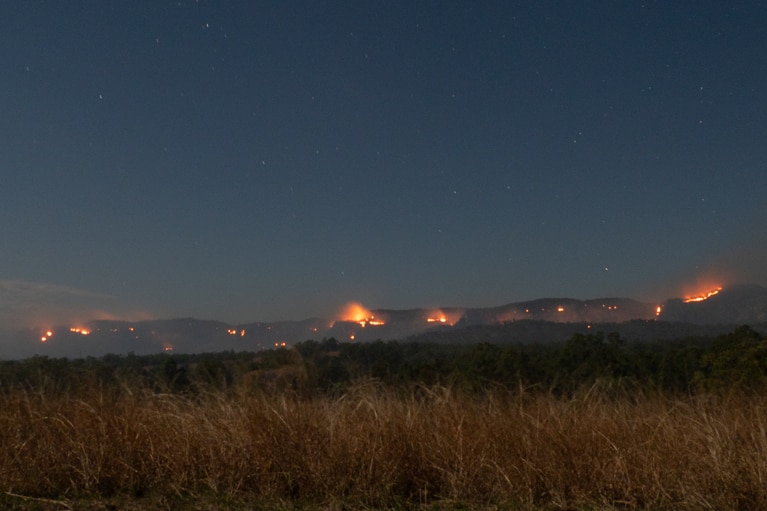Grass in foreground, sandstone cliffs, trees, multiple fires in background lighting up dark sky.