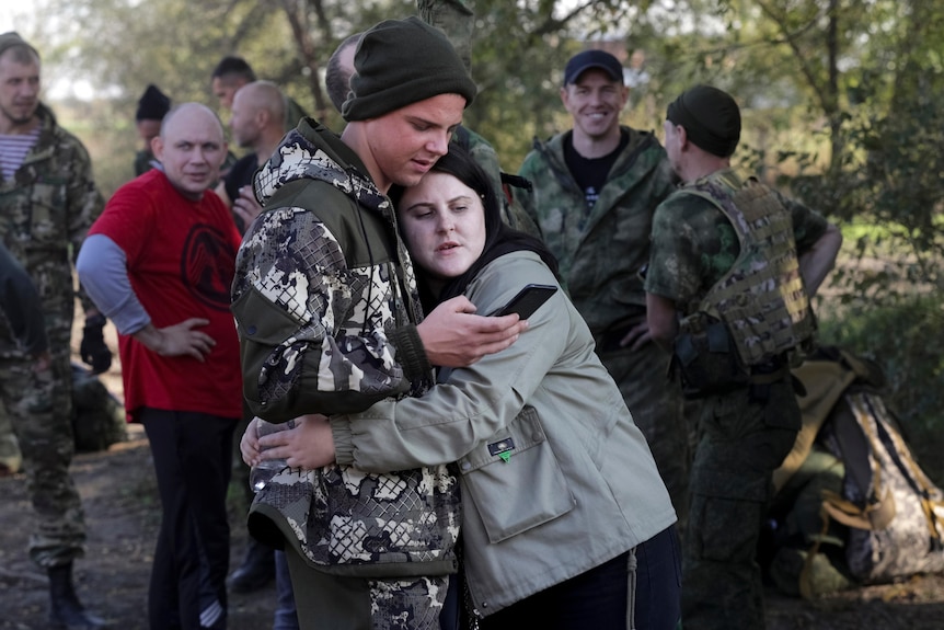 A woman and a man in uniform hug as they look at a black phone with other army recruits behind them