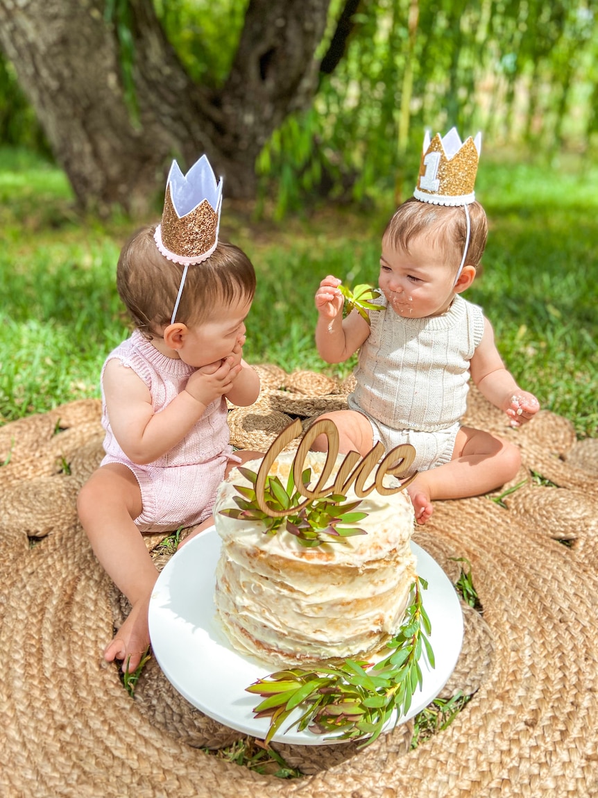 Twin babies Mia and Ava wearing golden party hats shaped like crowns, sitting in front of a big cake topped with the word "One".