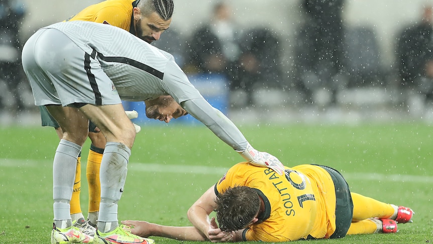 An injured Socceroos player lies on the pitch in the rain as a goalkeeper pats him on the back.