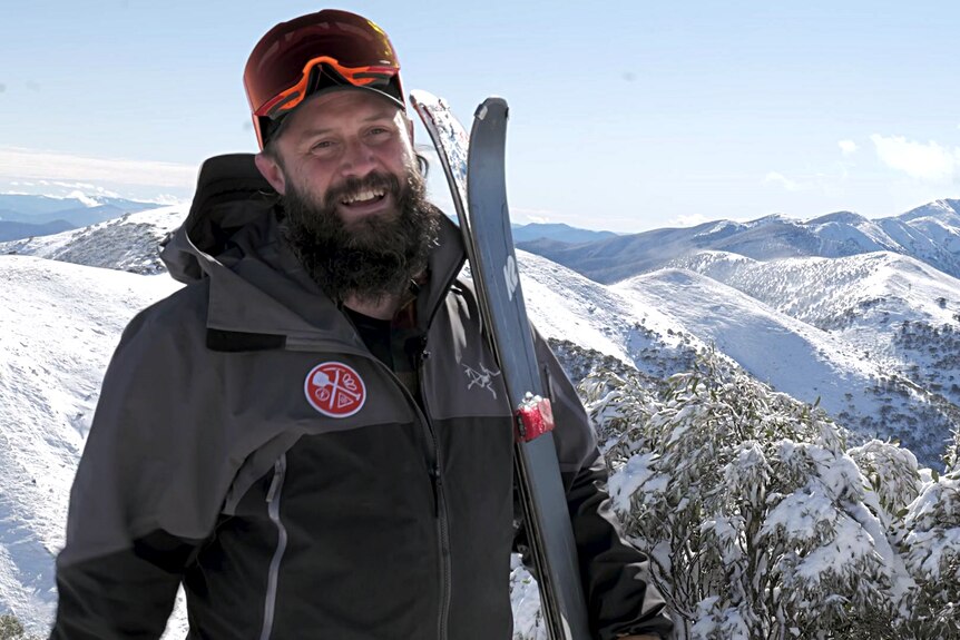 A man with a beard rests his skis against his shoulder.