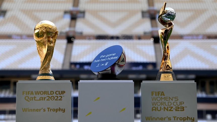 Two football World Cup trophies are seen on pedestals in a stadium, with a half-football on a pedestal between them.