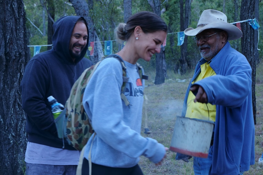 An older Indigenous man swings a can of smoke as two hikers smile and walk past.