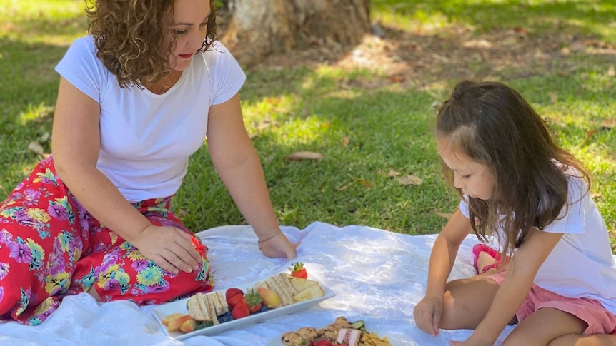 Mum Tara Leong sits on the ground looking at picnic sandwiches and strawberries with six year-old daughter