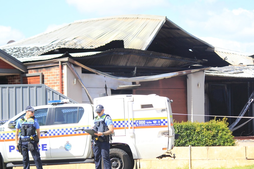 Two police officers in front of a burnt-out house. There is a police van parked outside.