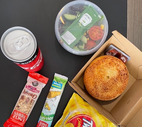 A can of soft drink, burger, salad in a plastic container, and a muesli bar and fruit roll up on a table