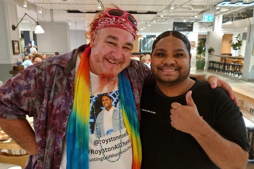 A man with a dyed beard, wearing a bandana, stands next to a smiling man giving a thump up.