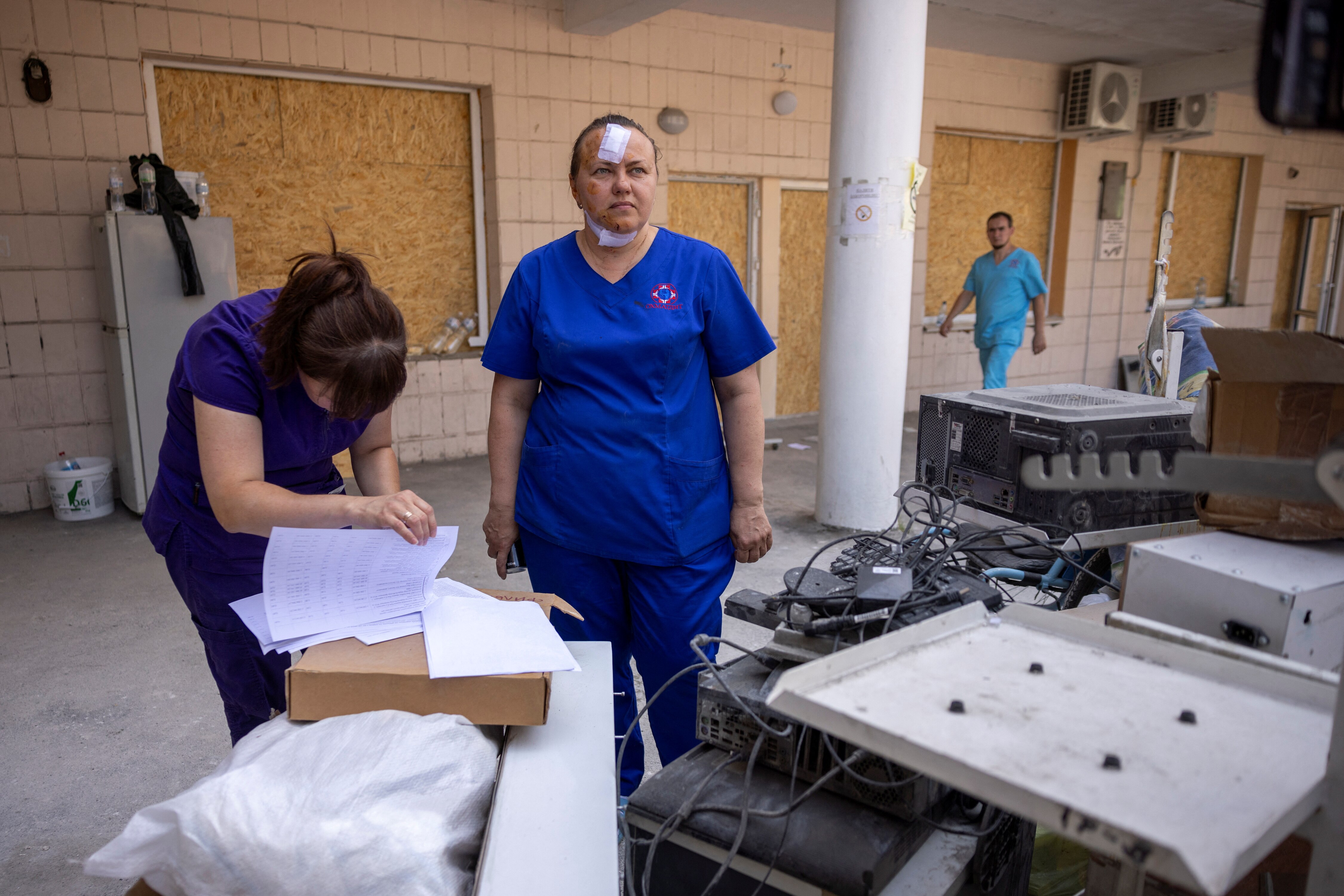 Two nurses in blue scrubs stand among medical equipment with boarded up windows behind them