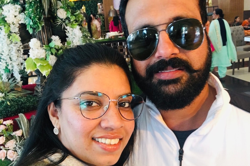 woman with glasses and black hair half tied back smiles next to a man with aviator sunglasses, beard, goatee and white polo