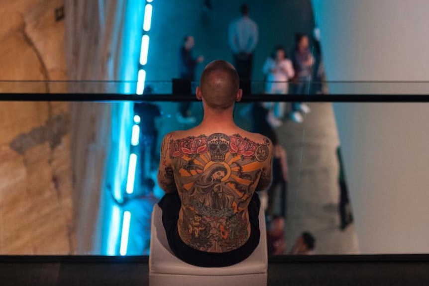Tattooed man looks down on people on a walkway below.