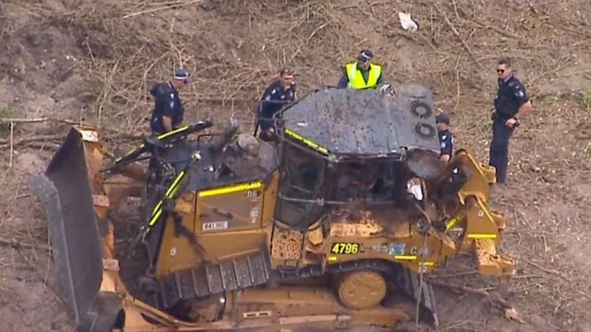 Several police officers stand around a bulldozer on a rural property