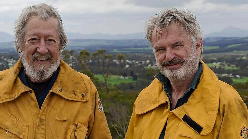 Two grey-haird, bearded men stand before a green countryside in yellow fire fighting uniforms.
