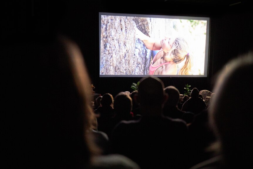 People in a dark cinema watch a screen showing a woman with a ponytail climbing a boulder.