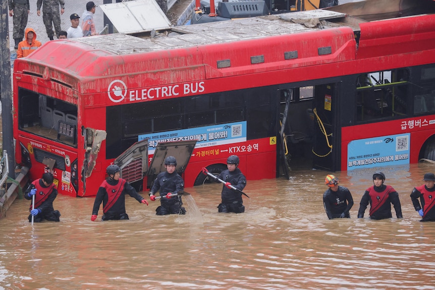 Rescuers wade waist-deep in floodwaters in front of a submerged bus. 