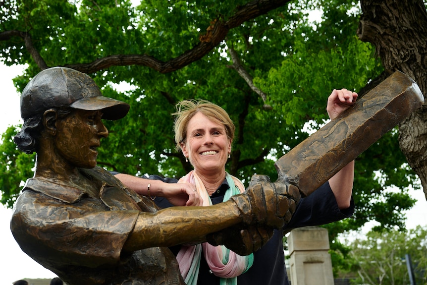 Australian cricket great Belinda Clark smiles as she stands behind a bronze statue of her playing an attacking shot.