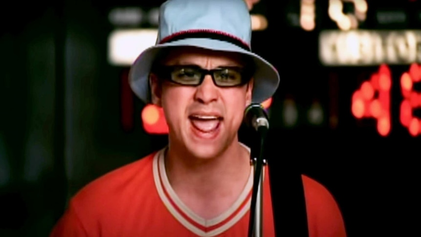 Singer of Wheatus wearing a blue bucket hat singing and playing guitar on a basketball court