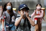 A small Asian boy in blue cap touches his face mask as he walks outside with two masked girls behind