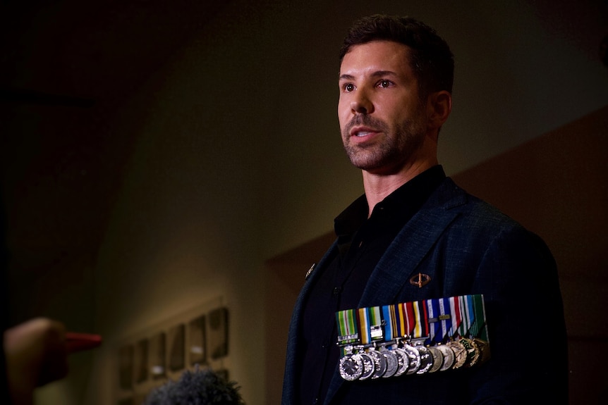 A man with short dark hair and military medals pinned to his chest speaks to the media. He wears a serious facial expression.