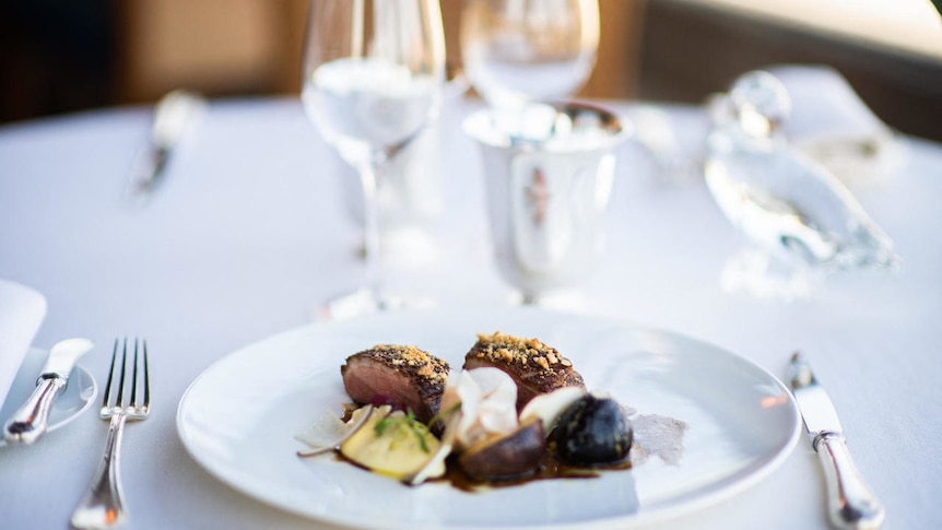 A fine-dining plate of meat and vegetables on a white tablelcloth with sparkling cutlery.