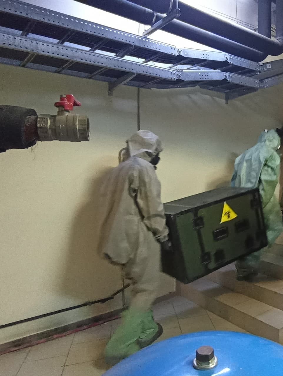 Two people in hazmat suits carrying a trunk with a biohazard symbol