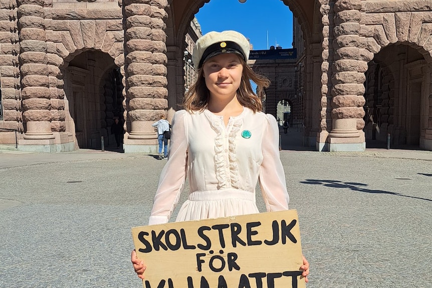 a young woman in a white dress and white cap holds a sign in front of parliament
