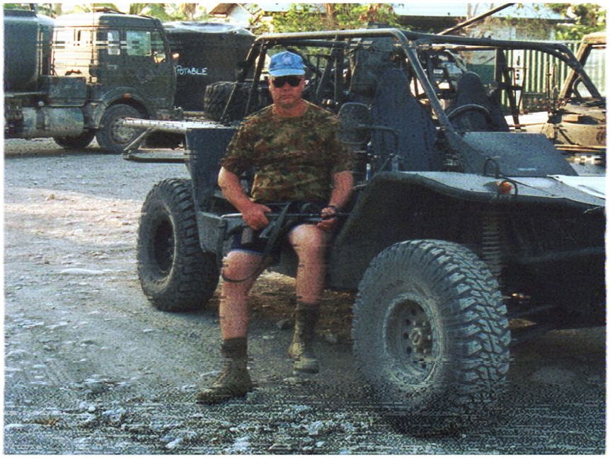 A man wearing a camouflage shirt and with a rifle on his lap, sits on the side of a buggy.