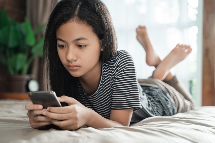 A young girl of Asian heritage is on a bed, looking at a smartphone