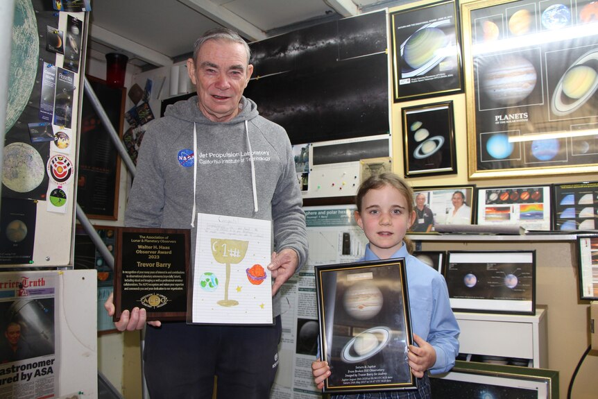 An older man and young girl holding certificates in a brightly lit room decorated with planetary pictures