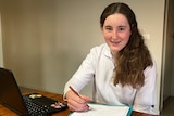 A teenage girl with long brown hair sits at a table, holds and pen and smiles.