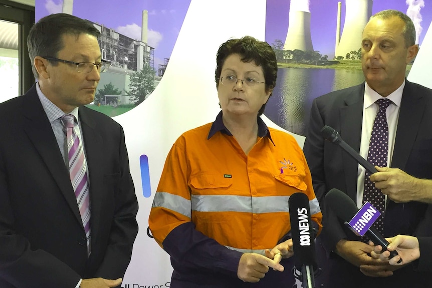 Two men in suits flank a woman in high-vis clothing who is standing in front of a microphone.