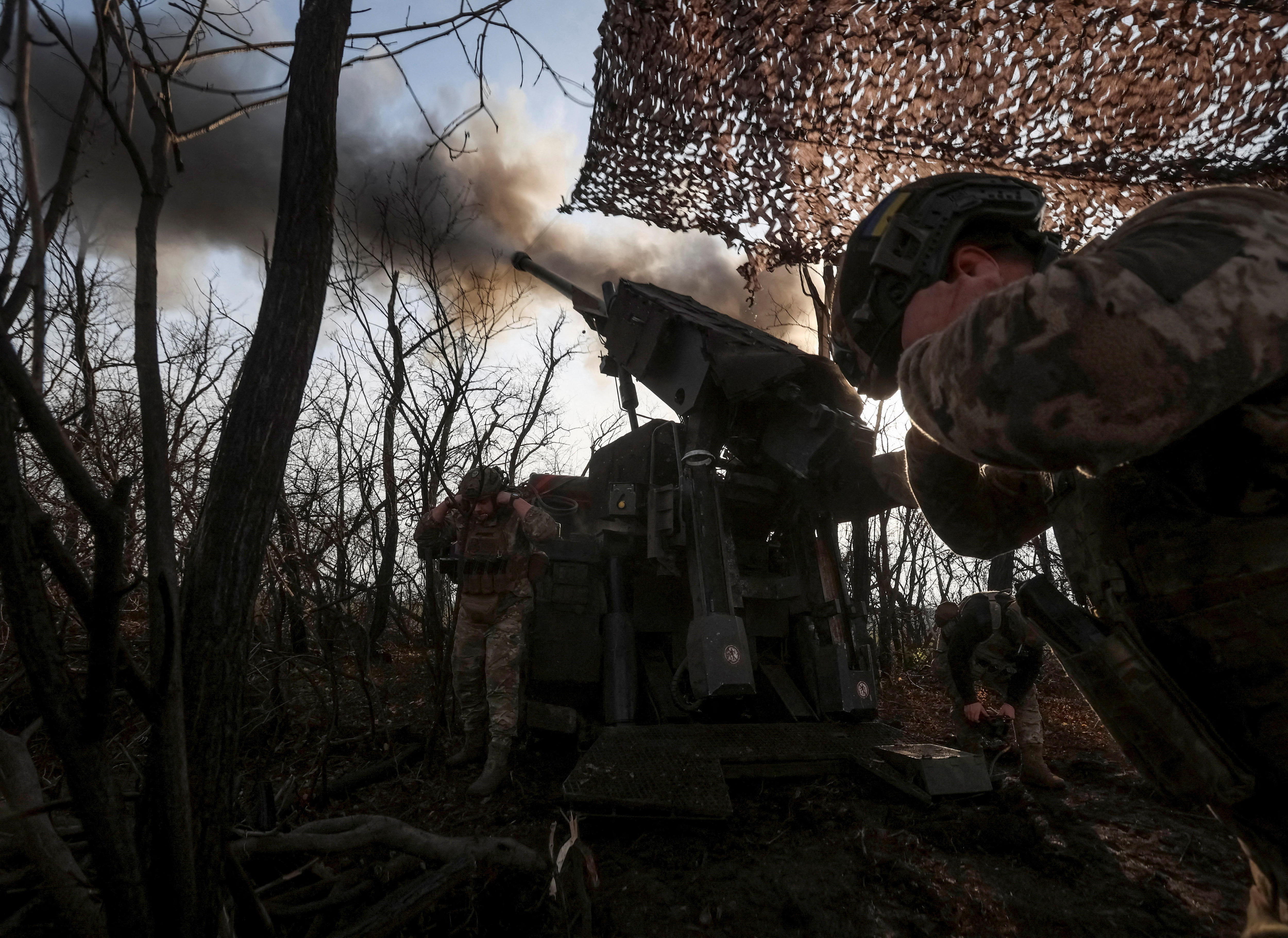 Two Ukrainian soldiers holding their ears as a Howitzer gun fires a mortar round in the middle of a forest.