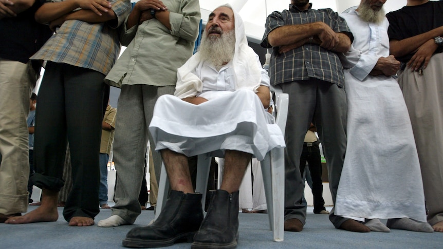Man with long beard wearing white clothes and boots sits on a chair. Men stand either side of him.