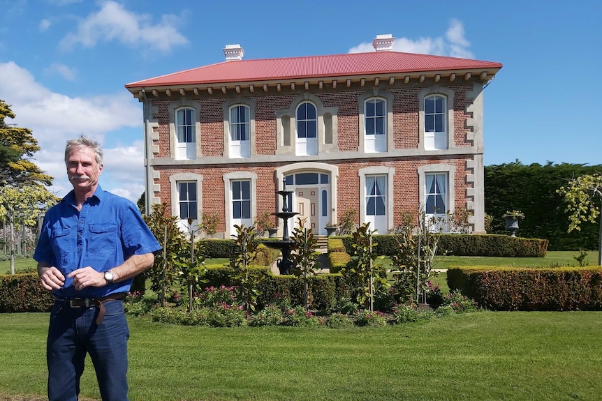 A man stands in front of a double-storey Georgian Homestead.
