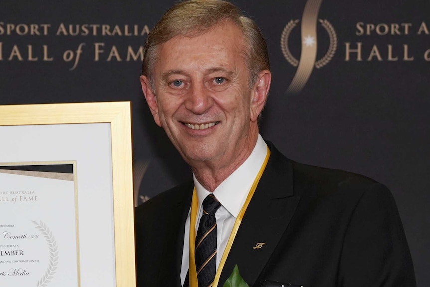 Dennis Cometti, with a medal around his neck, smiles while holding a framed certificate